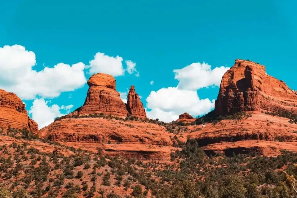 Geschichtete rote Felsformationen erheben sich aus einer Landschaft grüner Sträucher unter einem strahlend blauen Himmel mit bauschigen weißen Wolken.