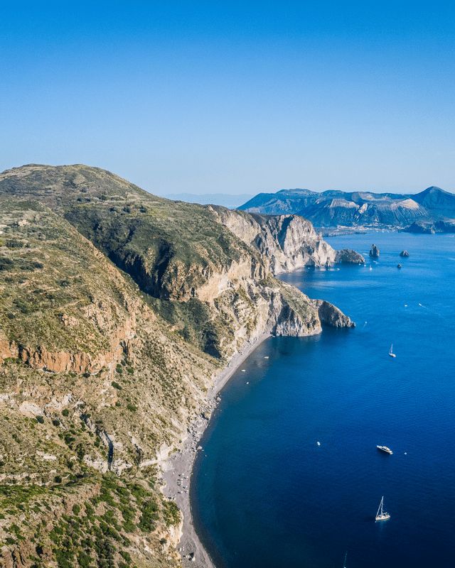 Vue aérienne d'un littoral accidenté et verdoyant rencontrant une mer d'un bleu profond, avec une plage de galets sombres et plusieurs voiliers dans la baie.