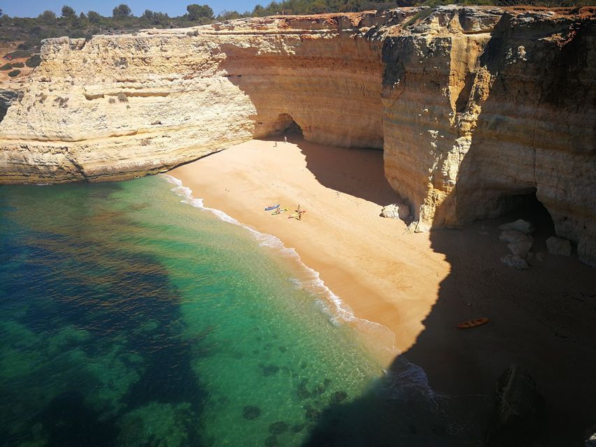 Ein abgelegener Sandstrand mit klarem, türkisfarbenem Wasser, umgeben von hohen Klippen mit Höhlen, aus der Vogelperspektive gesehen.