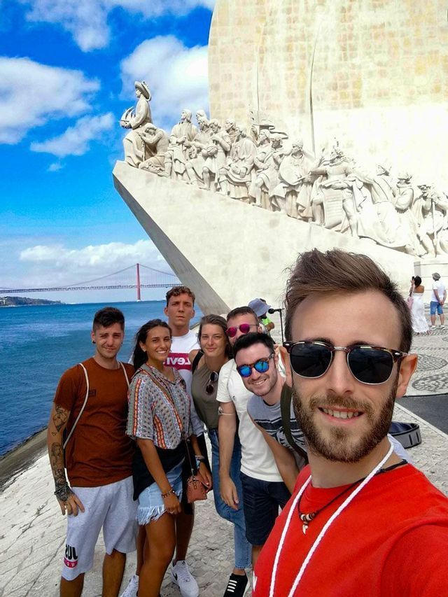 Eine WeRoad-Gruppe macht ein Selfie vor einem großen Steinmonument mit Brücke und Wasser im Hintergrund.