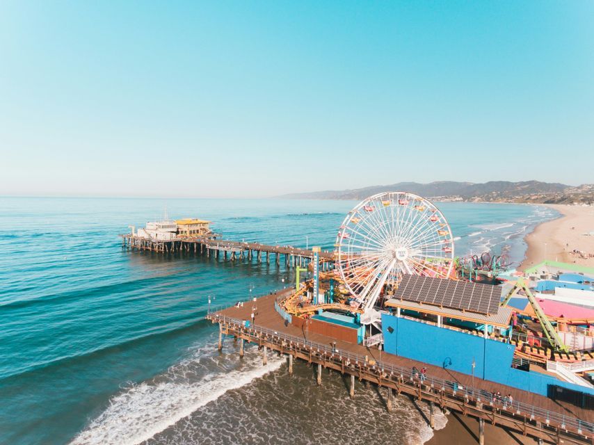 An aerial view of an amusement park pier with a large Ferris wheel over the ocean, next to a sandy beach under a clear sky.