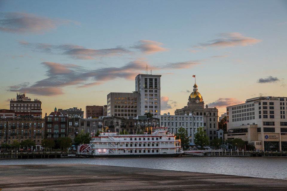 The Georgia Queen riverboat is docked on a river in front of a city skyline with a golden dome building at sunset.