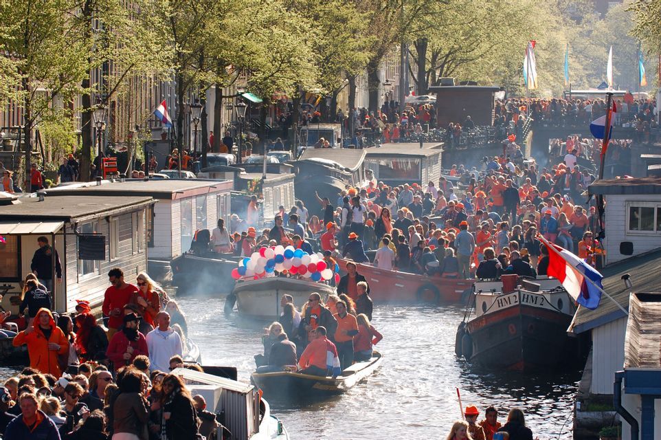 Una grande folla di persone festeggia su barche durante un festival lungo un trafficato canale cittadino fiancheggiato da case galleggianti.