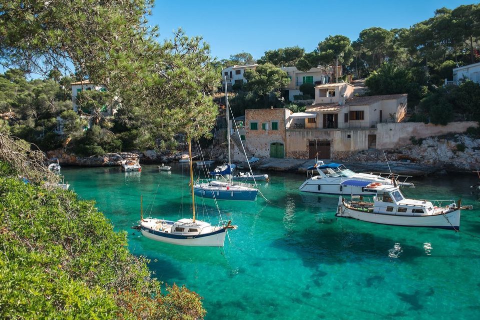 Diverse barche sono ancorate in una baia calma e turchese, circondata da alberi rigogliosi e case su una costa rocciosa.