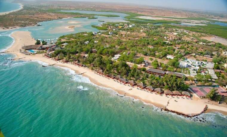 Una vista aerea di una costa tropicale con una spiaggia di sabbia, acqua turchese e un resort immerso tra alberi verdi e una laguna.