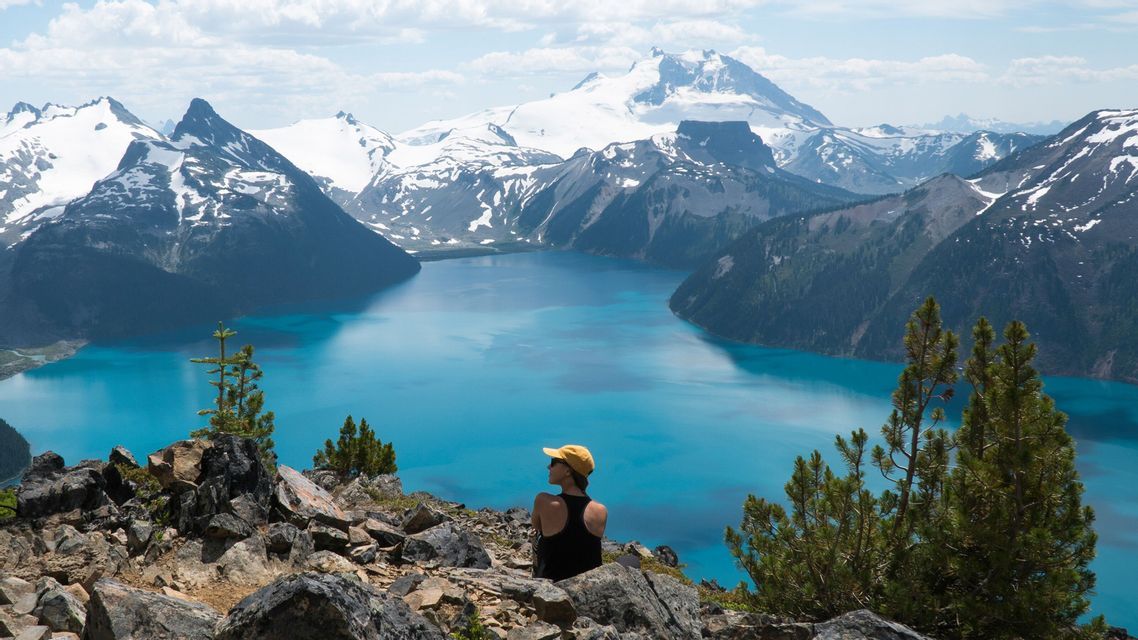 A woman in a yellow cap sits on a rocky peak, looking out over a turquoise alpine lake surrounded by snow-capped mountains.