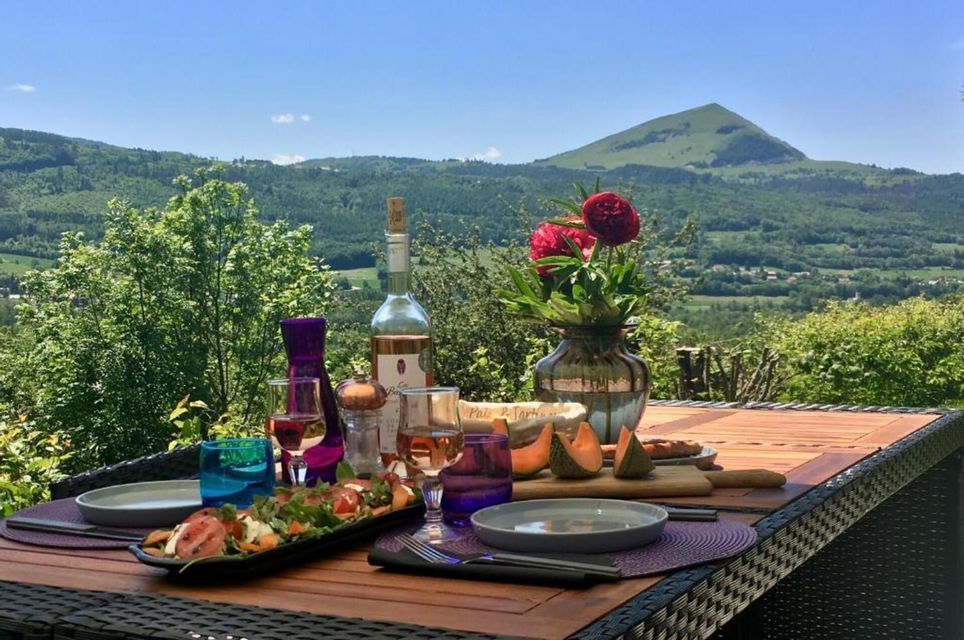 Une table dressée avec vin, fromage et salade sur une terrasse avec vue panoramique sur des collines verdoyantes et une montagne.