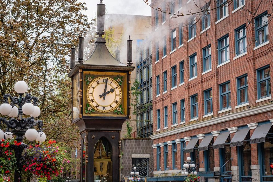 An ornate steam clock releases steam from its pipes on a city street next to a red brick building.