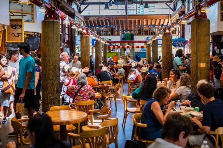 A bustling indoor food hall filled with people eating at wooden tables and walking around under a high ceiling with wooden beams.