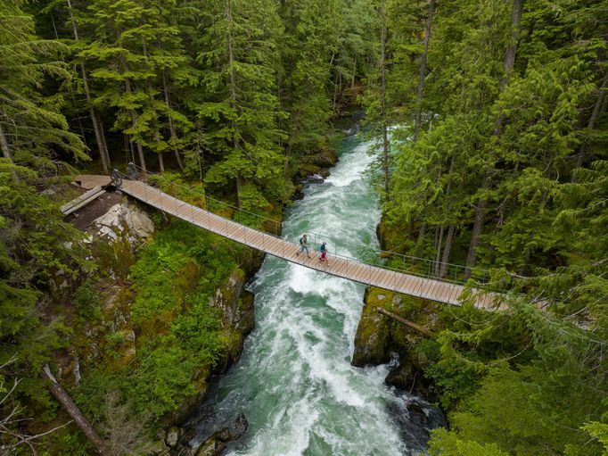 An aerial view of two people walking on a suspension bridge over a rushing river, surrounded by a dense green forest.