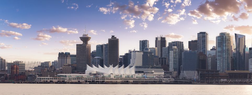 A panoramic view of a modern city skyline across the water, with skyscrapers and unique architecture under a cloudy sky at sunset.