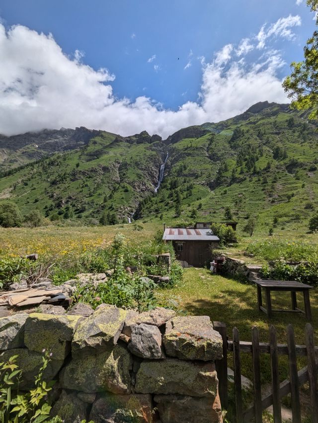 Un chalet rustique dans une prairie de montagne verdoyante avec une cascade lointaine, vu par-dessus un mur de pierre au premier plan.