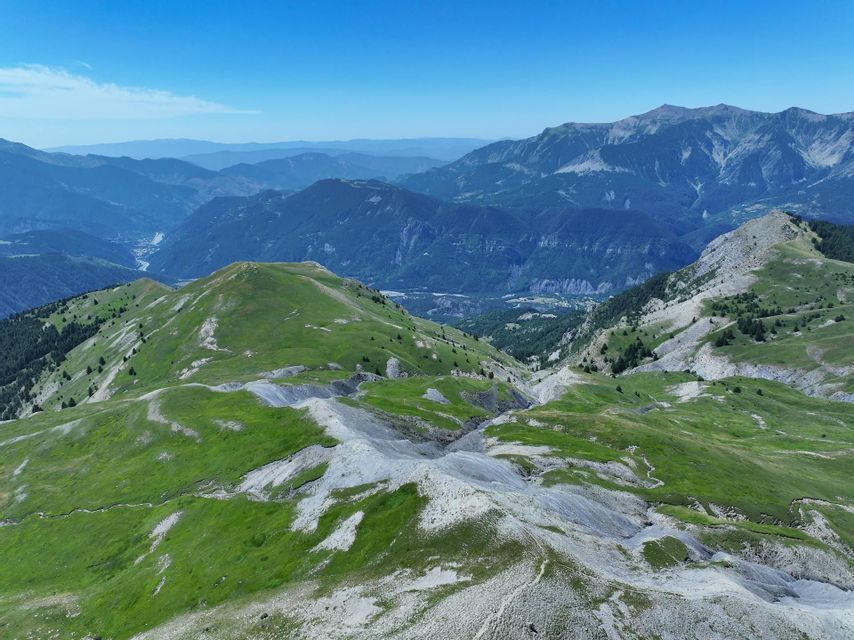 Vue aérienne d'une vaste chaîne de montagnes avec des collines verdoyantes et des pentes rocheuses sous un ciel bleu clair.