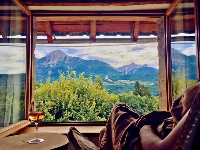 Une personne se détend avec un verre de vin devant une fenêtre ouverte, contemplant une vallée verdoyante et une chaîne de montagnes.