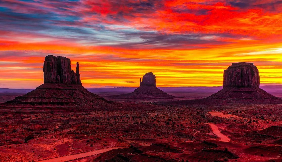 Three large rock buttes are silhouetted against a vibrant red and orange sunset over a desert landscape with a winding road.