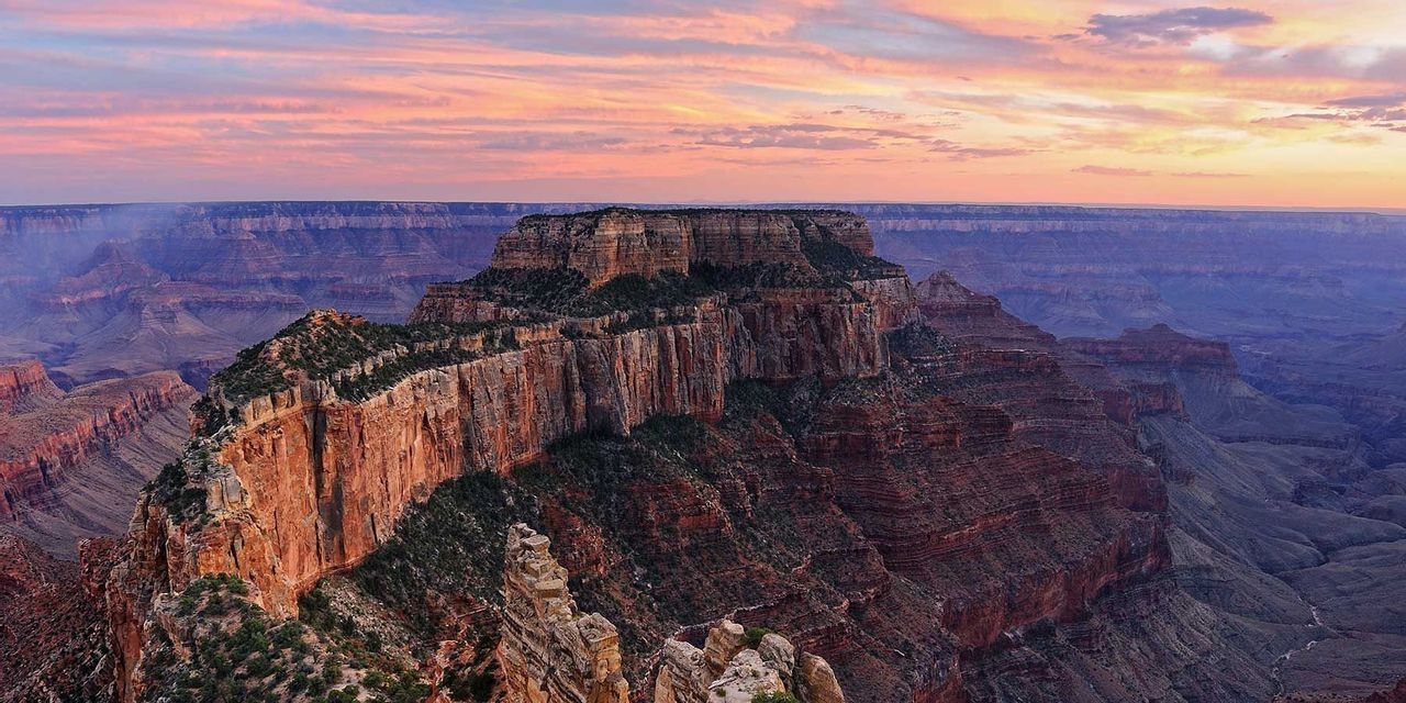 A panoramic view of a vast, layered canyon with a prominent mesa under a colorful pink and orange sunset sky.