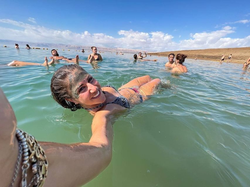 Una mujer sonriente con una mascarilla de barro en la cara flota en el agua, tomándose una selfie con su viaje en grupo de WeRoad de fondo.