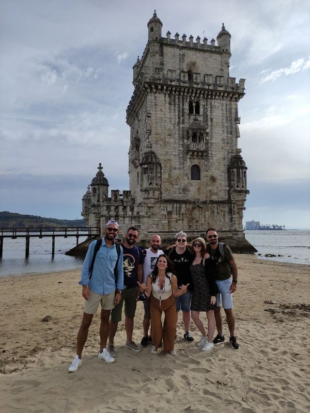 Un viaggio di gruppo WeRoad di sette persone in posa su una spiaggia sabbiosa di fronte a una grande torre di pietra in riva al mare sotto un cielo nuvoloso.