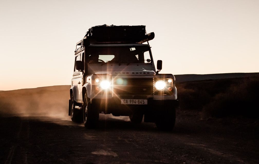 Ein weißer Land Rover mit einer WeRoad-Gruppenreise fährt bei Sonnenuntergang mit eingeschalteten Scheinwerfern auf einer staubigen Straße.