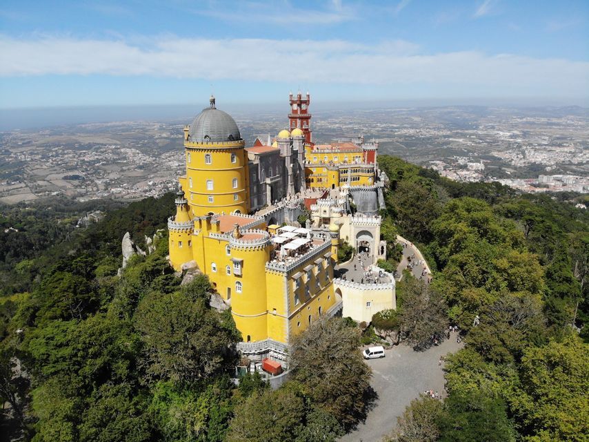 Un colorido castillo amarillo y rojo con torres se alza en una colina boscosa, dominando una extensa ciudad bajo un cielo azul.