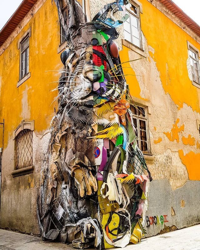 En la esquina de un edificio amarillo envejecido, se encuentra una gran escultura de un conejo hecha con materiales reciclados de colores.