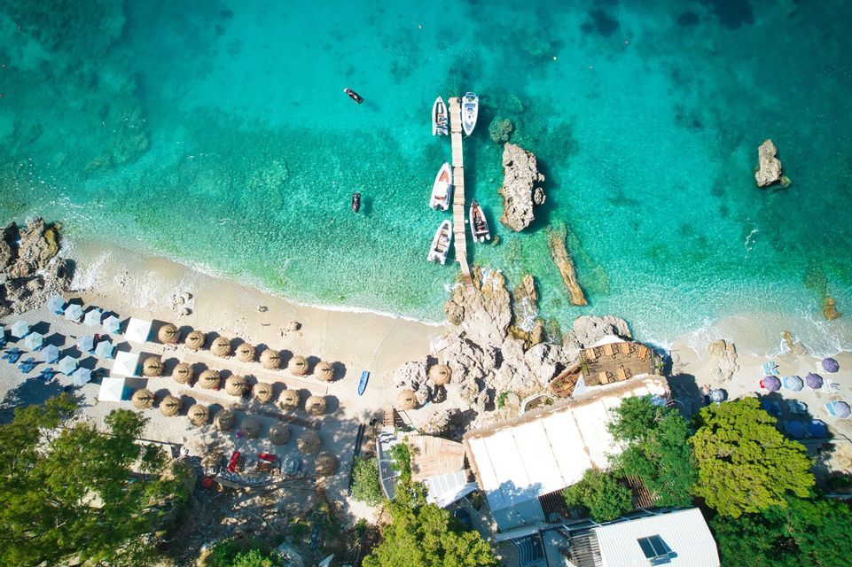 Luftaufnahme eines sonnigen Strandes mit klarem türkisen Wasser, einem Holzsteg mit Booten und Reihen von Strohschirmen.
