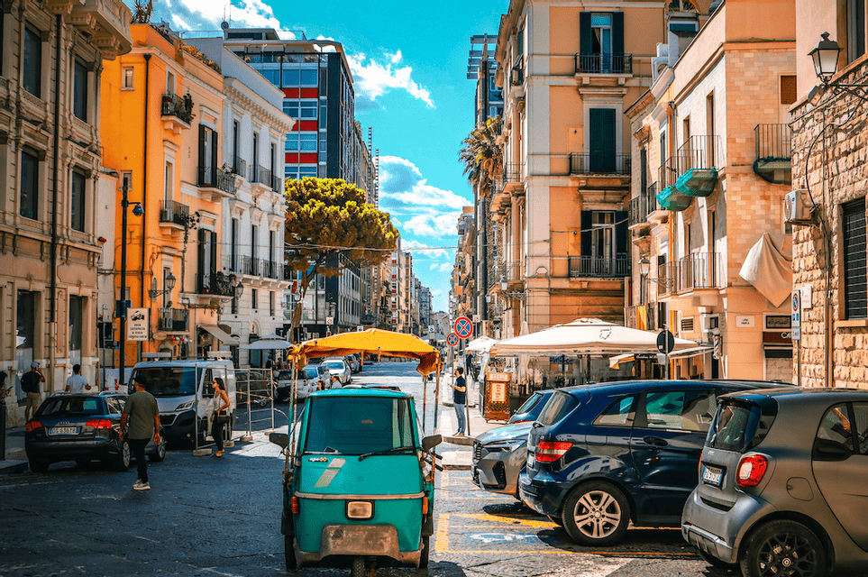Un veicolo verde a tre ruote parcheggiato su una strada cittadina fiancheggiata da edifici colorati e altre auto sotto un cielo azzurro brillante.