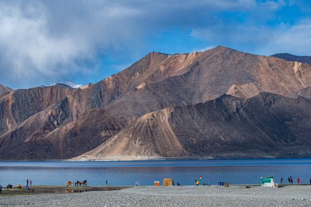 Un vasto lago azul se asienta en la base de grandes y escarpadas montañas, con personas y animales en la orilla rocosa.