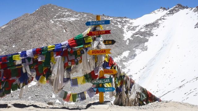 Un cartel adornado con coloridas banderas de oración indica direcciones a lugares como Siachen, Leh y Kargil en un puerto de montaña nevado.