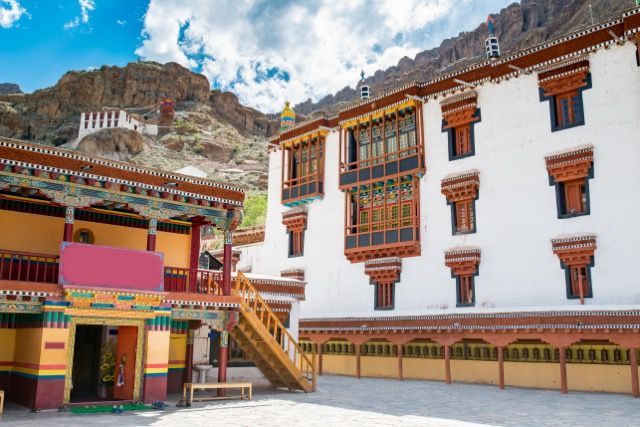 Un monasterio tradicional y ornamentado, con muros blancos y ventanas de madera de colores, se erige en un patio al pie de una montaña rocosa.