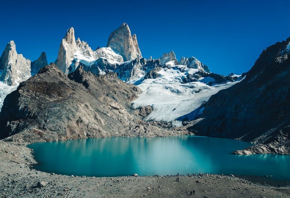 Un groupe WeRoad longe la rive rocheuse d'un lac turquoise, au pied de montagnes déchiquetées et enneigées, sous un ciel bleu clair.