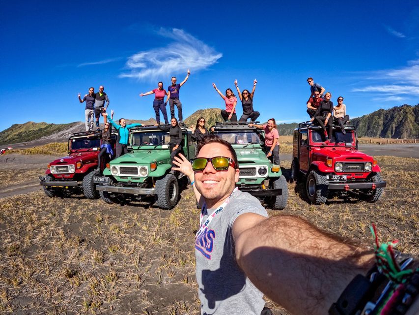 Un hombre se toma una selfie con un grupo de WeRoad posando sobre y alrededor de coloridos jeeps todoterreno en un vasto paisaje árido bajo un cielo azul.