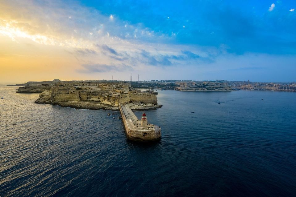 Vista aerea di un frangiflutti in pietra e un faro che si estendono da una città costiera fortificata verso il mare al tramonto.