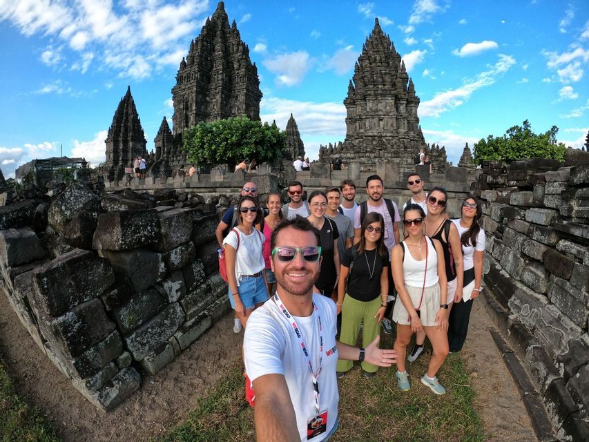 Un viaje en grupo de WeRoad se toma una selfie de gran angular frente a un antiguo complejo de templos de piedra bajo un cielo parcialmente nublado.
