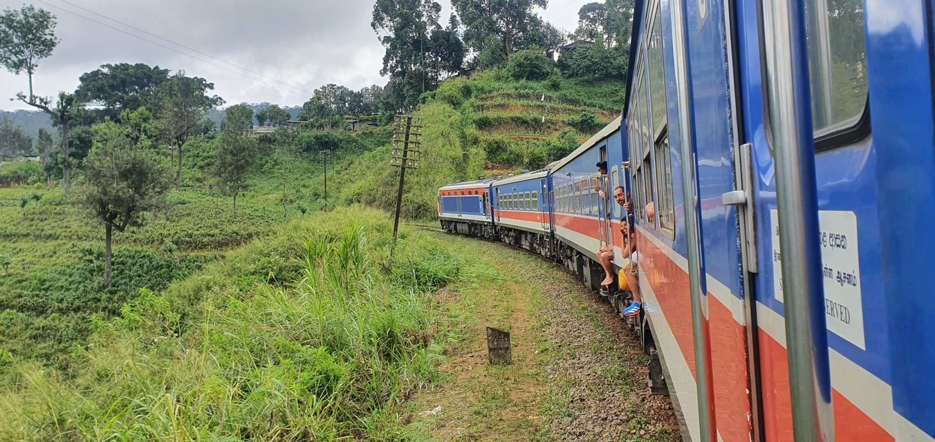 Un grupo de WeRoad viaja en las puertas abiertas de un tren azul y rojo a través de un paisaje exuberante, verde y montañoso.