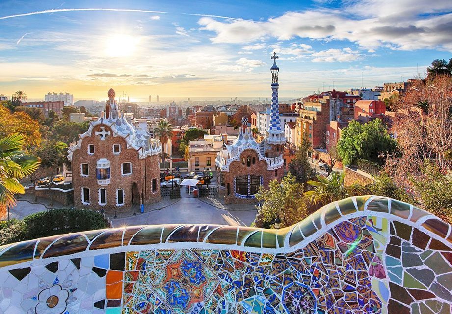 Una panchina a mosaico colorata si affaccia su due edifici stravaganti e su una città che si estende fino al mare sotto un cielo soleggiato.