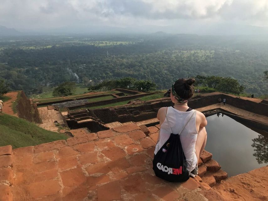 A person with a WeRoad backpack sits on brick ruins overlooking a vast, forested valley from a high viewpoint.