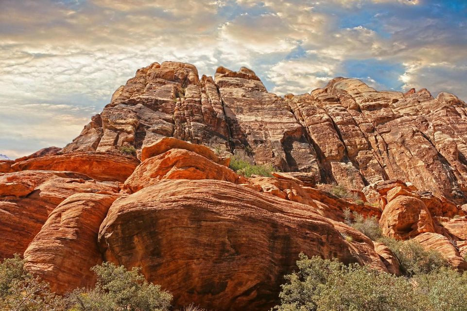 A large, layered red rock formation rises towards a partly cloudy sky, with green shrubs visible at its base.