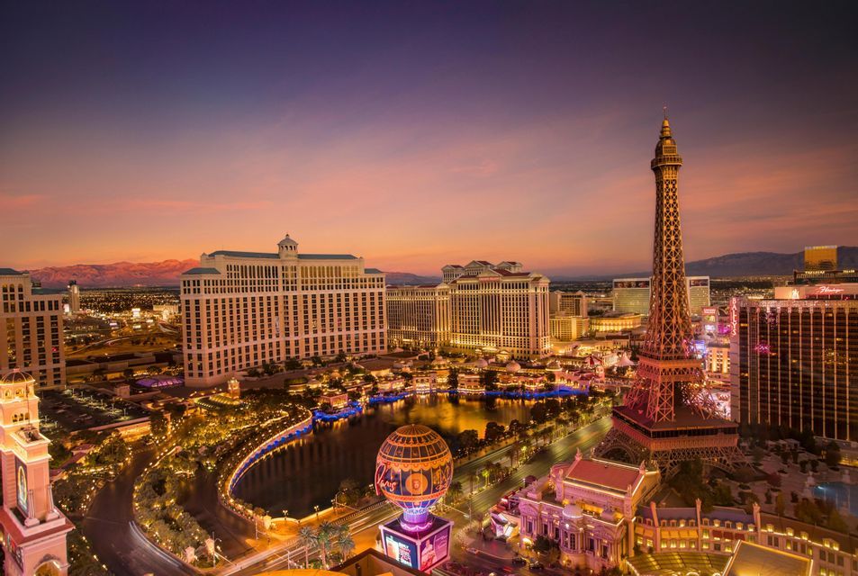 An aerial view of an illuminated cityscape featuring a replica Eiffel Tower and large hotels under a colorful sunset sky.