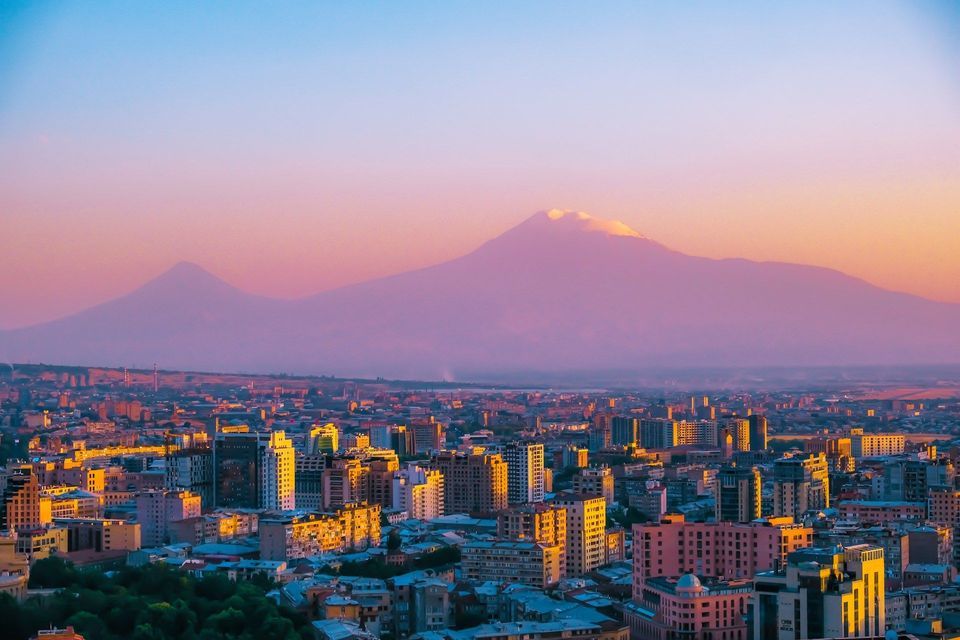 Un paysage urbain dense, avec des bâtiments éclairés par le coucher de soleil et une grande montagne à double sommet en arrière-plan sous un ciel coloré.