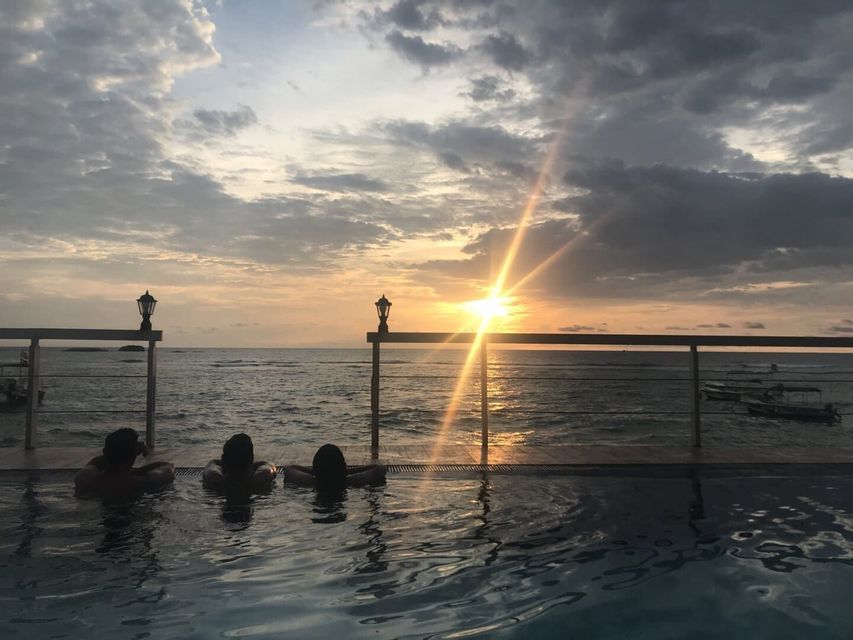 Drei Personen einer WeRoad Gruppenreise entspannen in einem Infinity-Pool mit Blick auf das Meer während eines bewölkten Sonnenuntergangs.