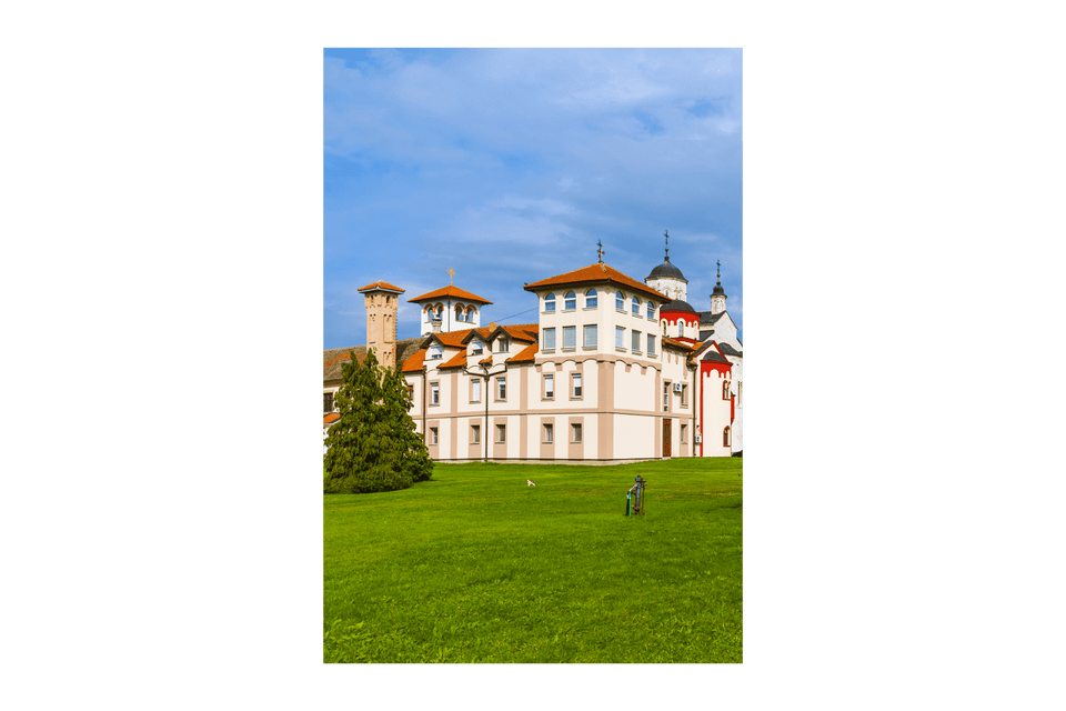 Un grande monastero con tetti di tegole rosse e cupole ortodosse si erge dietro un vasto prato verde sotto un cielo azzurro con nuvole leggere.
