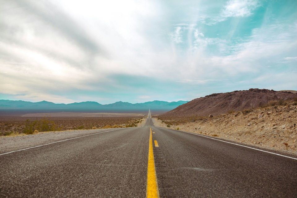 A long, straight asphalt road with a yellow line leads through a vast desert landscape toward distant mountains.
