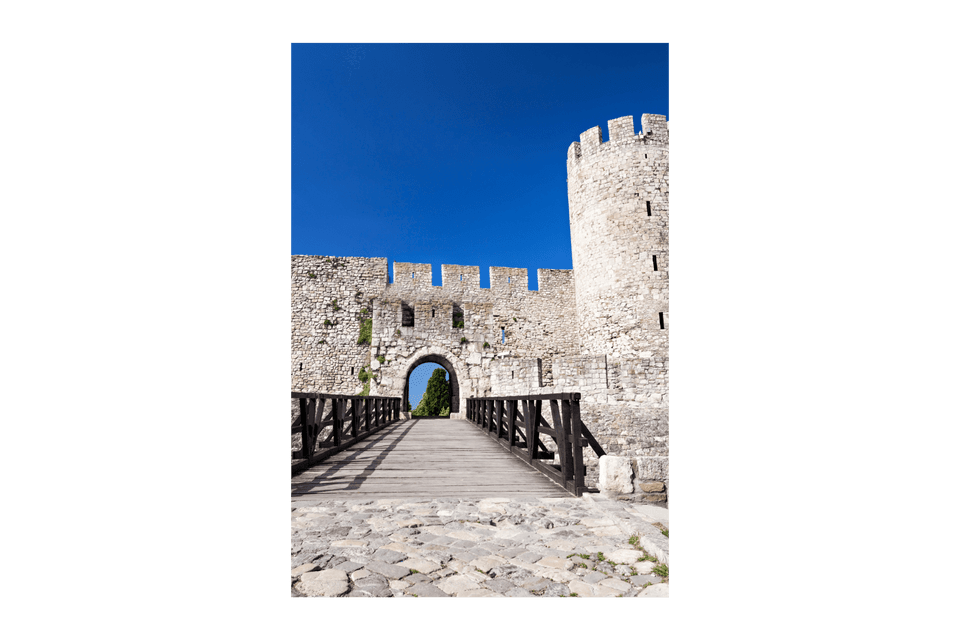 Un ponte di legno conduce all'ingresso ad arco di una fortezza in pietra con mura merlate e una torre, sotto un cielo azzurro e limpido.