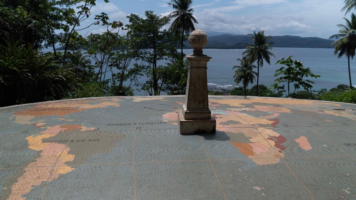 Un monumento in pietra su una grande mappa mondiale a mosaico che mostra l'America del Sud e l'Oceano Atlantico, con vista su una costa tropicale.