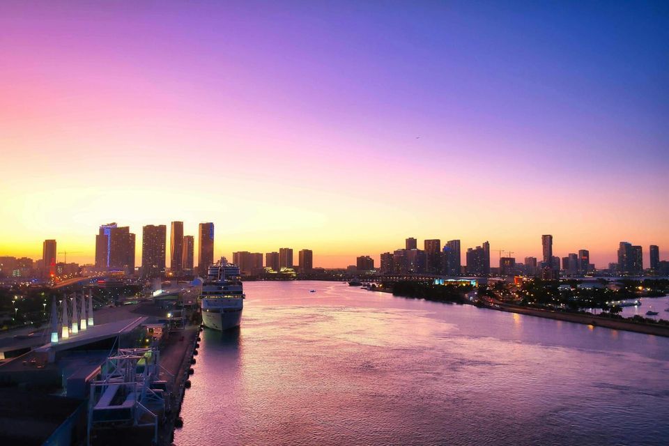 A city skyline along a wide waterway with a docked cruise ship, viewed under a purple and orange sunset.