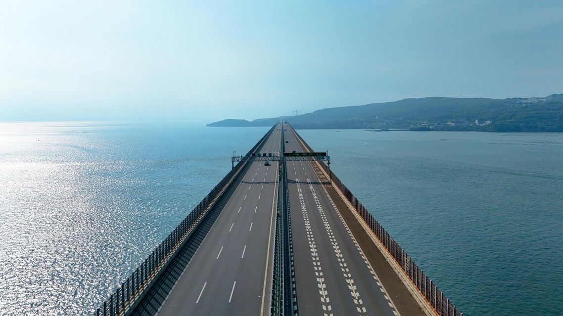 An aerial view of a long highway bridge stretching across the sparkling sea towards a distant, hilly coastline.