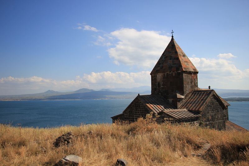 Une vieille église en pierre au toit de tuiles se dresse sur une colline herbeuse, dominant un grand lac avec des montagnes en arrière-plan.