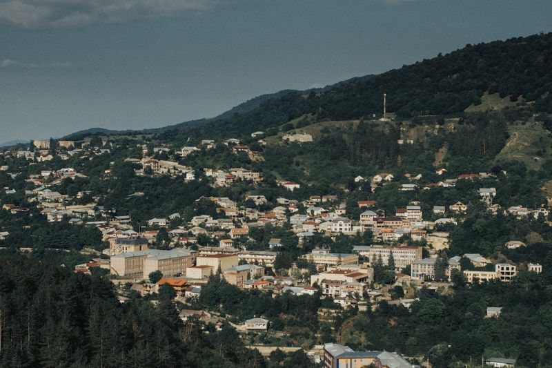 Une vue plongeante sur une ville avec des bâtiments dispersés sur une colline escarpée et boisée, sous un ciel partiellement nuageux.
