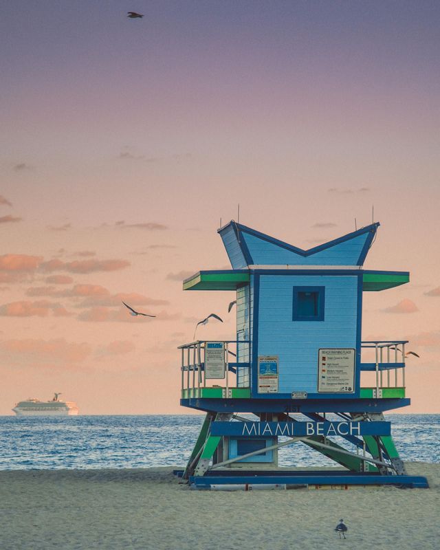 A blue and green lifeguard tower on Miami Beach at sunset, with a cruise ship on the ocean and seagulls flying by.
